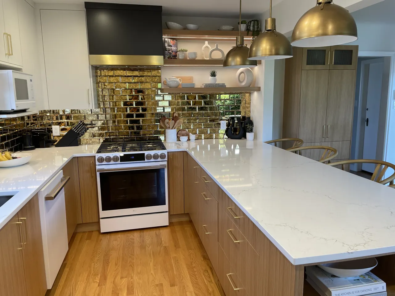 Finished kitchen renovation with wood-grain lower cabinets, white quartz peninsula, brass pendant lights and a mirrored gold tile backsplash