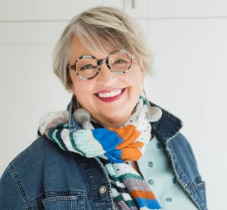 Sherry Willing, smiling, wearing round tortoiseshell glasses, a denim jacket and a colourful striped scarf